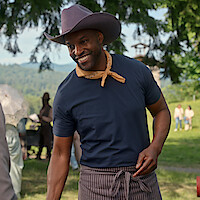 Smiling man in a cowboy hat, scarf, and apron outdoors at a sunny park or rural event, with people and trees in the background.