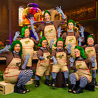 Group of people in Oompa Loompa costumes with green wigs, brown shirts, striped tights, and tan overalls pose and wave inside a colorful candy factory set with oversized sweets and whimsical machinery in the background.