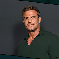 Alan Ritchson with short hair wearing a dark green collared shirt standing against a plain dark background, facing forward and smiling slightly in a studio setting.