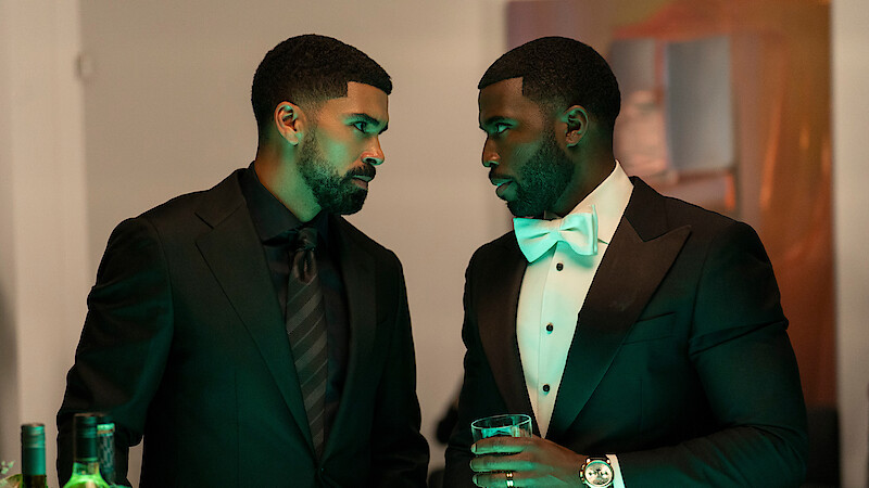 Two men in formal black suits stand close, engaged in a serious conversation at an indoor event. One holds a drink, bottles are visible on the table, and ambient green lighting enhances the elegant, upscale setting.