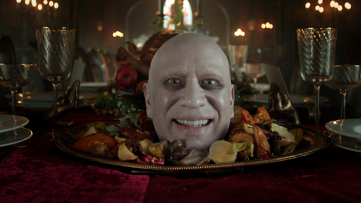 A smiling bald man’s head appears on a platter surrounded by roasted vegetables, set on an elegant dining table with ornate glasses and a dimly lit, candlelit gothic banquet hall in the background.