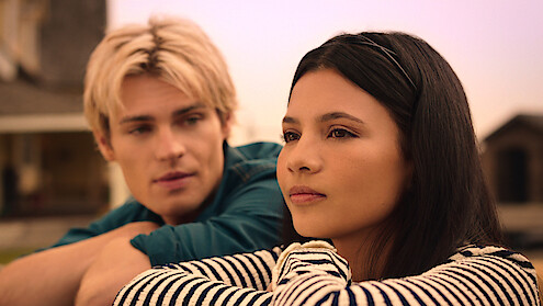 Two young people sit close outdoors at sunset, one with dark hair in focus looking thoughtful, and another with blond hair blurred in the background, both in casual clothes with a house structure visible behind them.