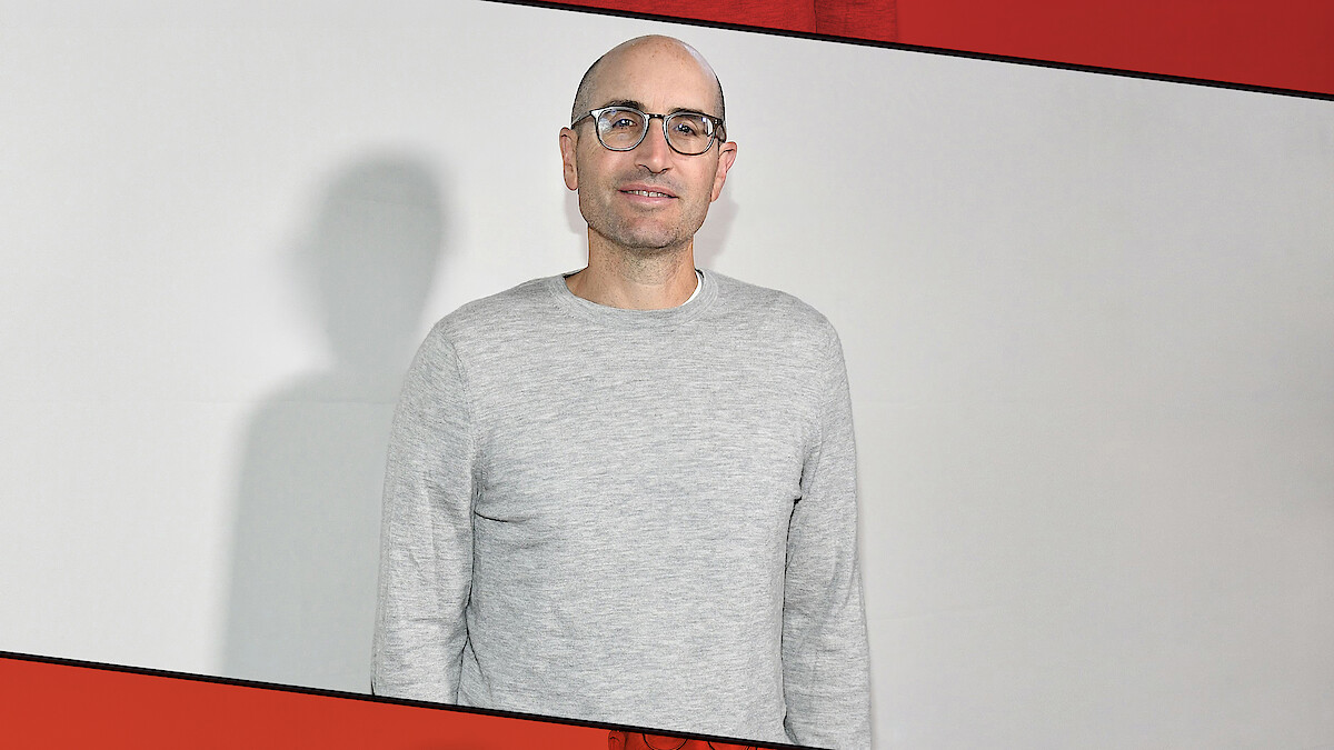 Man with glasses and a gray sweater stands in front of a plain white wall, casting a shadow, with a red and black diagonal border framing the image.