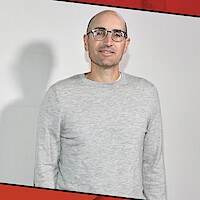 A man with glasses and a bald head wearing a light gray sweater stands indoors against a plain light-colored wall with a red border accent on the top and bottom edges.