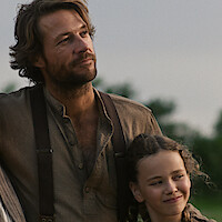 A man with tousled hair and a brown shirt stands outdoors with a young girl with braids; both look content, surrounded by greenery and soft natural light.