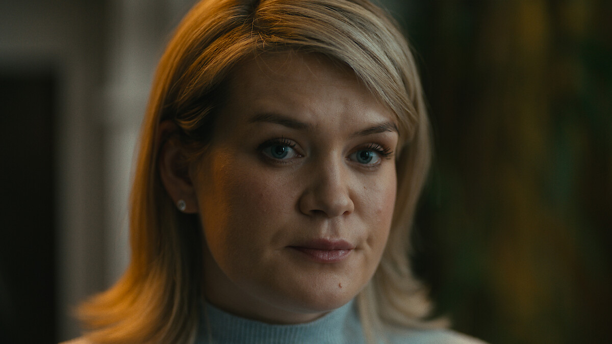 A woman with blonde hair in a blue turtleneck sweater sits indoors, softly lit, looking slightly serious with a blurred background of green and yellow tones.