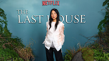 Greta Lee stands in front of a blue backdrop with "Netflix" and "The Last House" text, surrounded by greenery and natural elements, at a promotional event or premiere.