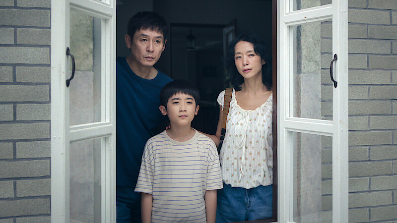 A man, woman, and young boy stand closely together inside a house, looking out through an open window with white frames. They appear serious, framed by gray brick walls and soft indoor lighting.