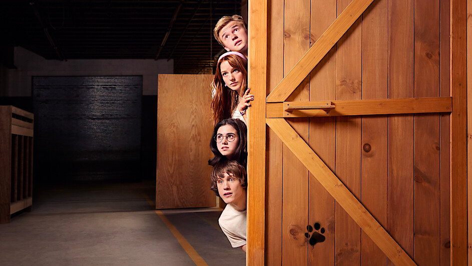 Four young people peeking around a wooden door with a paw print, in a dimly lit warehouse or storage environment, looking curious or cautious.
