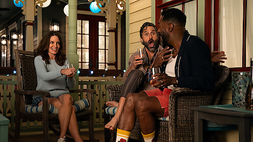 Three friends sit on a cozy porch in the evening, laughing and chatting while holding drinks, surrounded by warm lights and casual outdoor furniture.