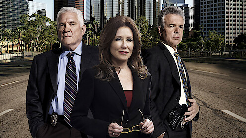 Three serious-looking detectives in suits stand on an empty city street with tall buildings and trees in the background, suggesting an urban law enforcement setting.