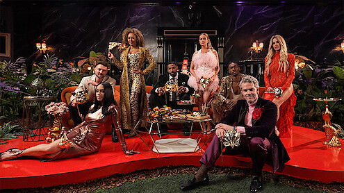 A group of elegantly dressed people holding masks pose in a lavish, dimly lit room with red carpets, gold decor, flowers, and lush plants, suggesting a masquerade party or formal event.