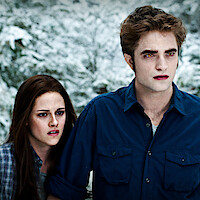 Kristen Stewart and Robert Pattinson stand close together in a snowy forest, both looking intensely ahead.