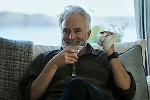 Bradley Whitford as Todd Penn in 'The Diplomat,' holds a martini glass in one hand and smiles while his other hand is wrapped in a cloth bandage.
