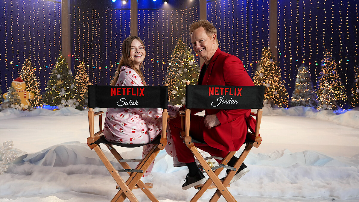 Two people smiling while sitting on director’s chairs with Netflix logos in a festive winter set decorated with snow, Christmas trees, twinkling lights, and holiday decorations, creating a cheerful holiday atmosphere.