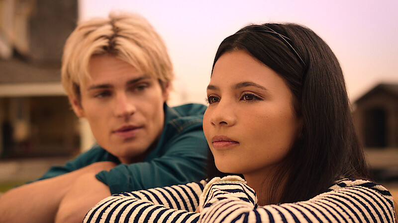 Two young people sit close outdoors at sunset, one with dark hair in focus looking thoughtful, and another with blond hair blurred in the background, both in casual clothes with a house structure visible behind them.