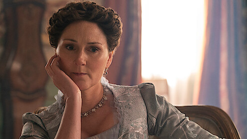A woman with styled brown hair and vintage clothing sits indoors on a chair, resting her head on her hand, with soft natural light and ornate décor in the background, evoking a historical or period setting.