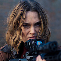Woman with determined expression aiming a rifle with a scope, outdoors against a blurred background, wearing a brown jacket, small red mark on forehead, intense focus in action scene setting.