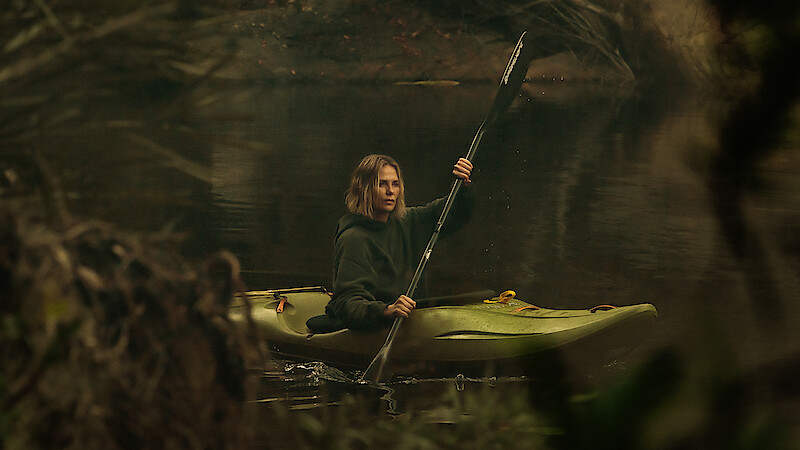 A person paddling a green kayak on a calm, forested river surrounded by trees and branches, wearing a dark hoodie and looking ahead with a serious expression, in a natural outdoor setting.