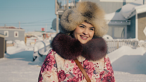 Smiling woman in a floral winter coat and fur hat standing outdoors in a snowy residential area, sunny day, snow-covered houses and fences in the background.