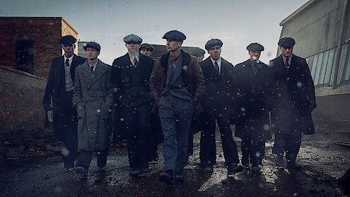 A group of men in vintage suits and newsboy caps walk together closely on a rainy, industrial street. The mood is serious and gritty, with cold light and falling rain enhancing the dramatic, early 20th-century atmosphere.