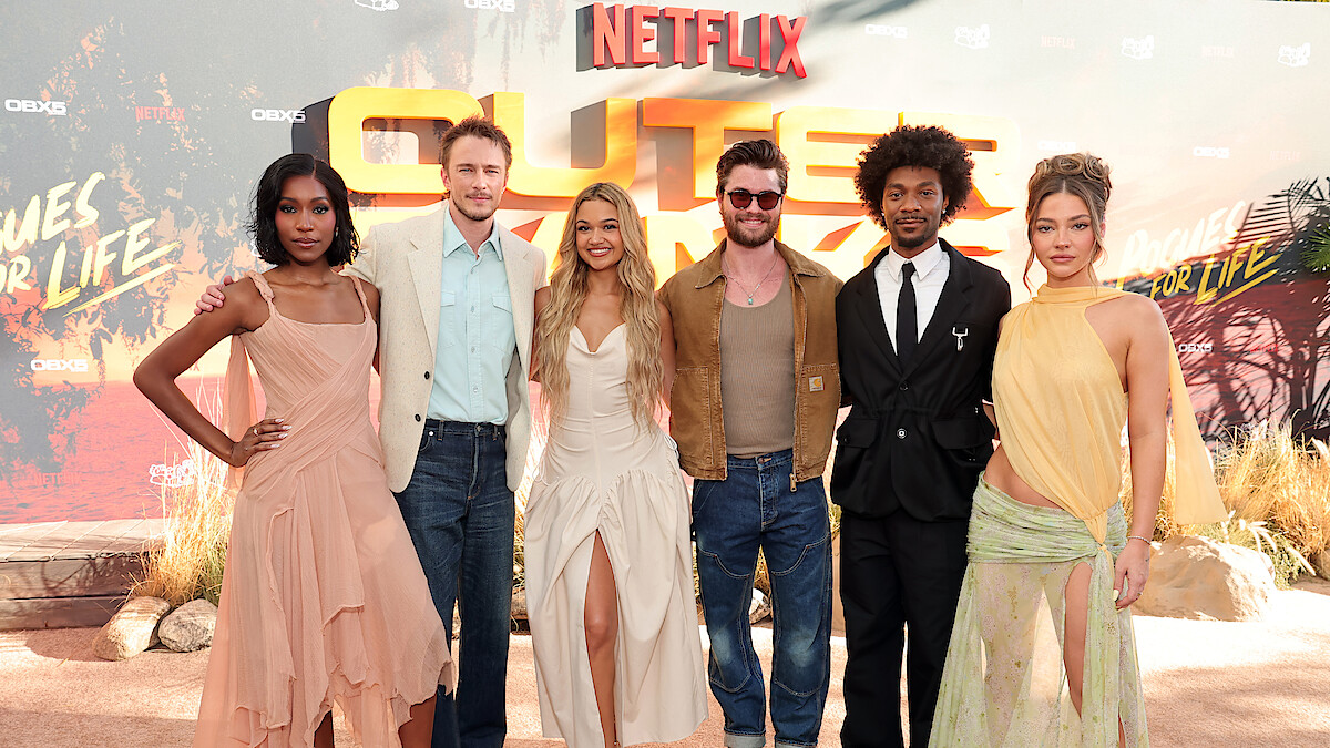 Six people pose together on a red carpet at an outdoor Netflix premiere event, dressed in fashionable attire, standing in front of a themed backdrop with desert elements and promotional text.