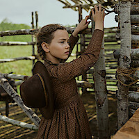 A young girl in a brown dress with a hat on her back stands inside a rustic wooden structure made of logs, looking thoughtfully towards the distance in a rural outdoor setting.