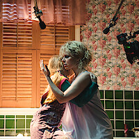 Woman hugs young girl in a vintage room with green tiles and floral wallpaper.
