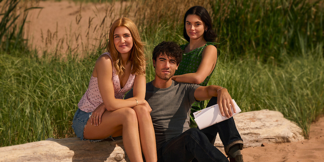 Three young adults sitting on rocks outdoors with tall green grass and sand in the background, enjoying a sunny day in a natural setting.