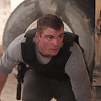 Man in tactical gear crouches indoors, appearing alert and focused, with a large tire and wooden structure in the background, possibly training or in a mission scenario.