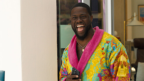 Man standing indoors near a doorway, laughing joyfully, wearing a colorful bathrobe with a bright pink collar and holding a smartphone, with modern home furnishings in the background.