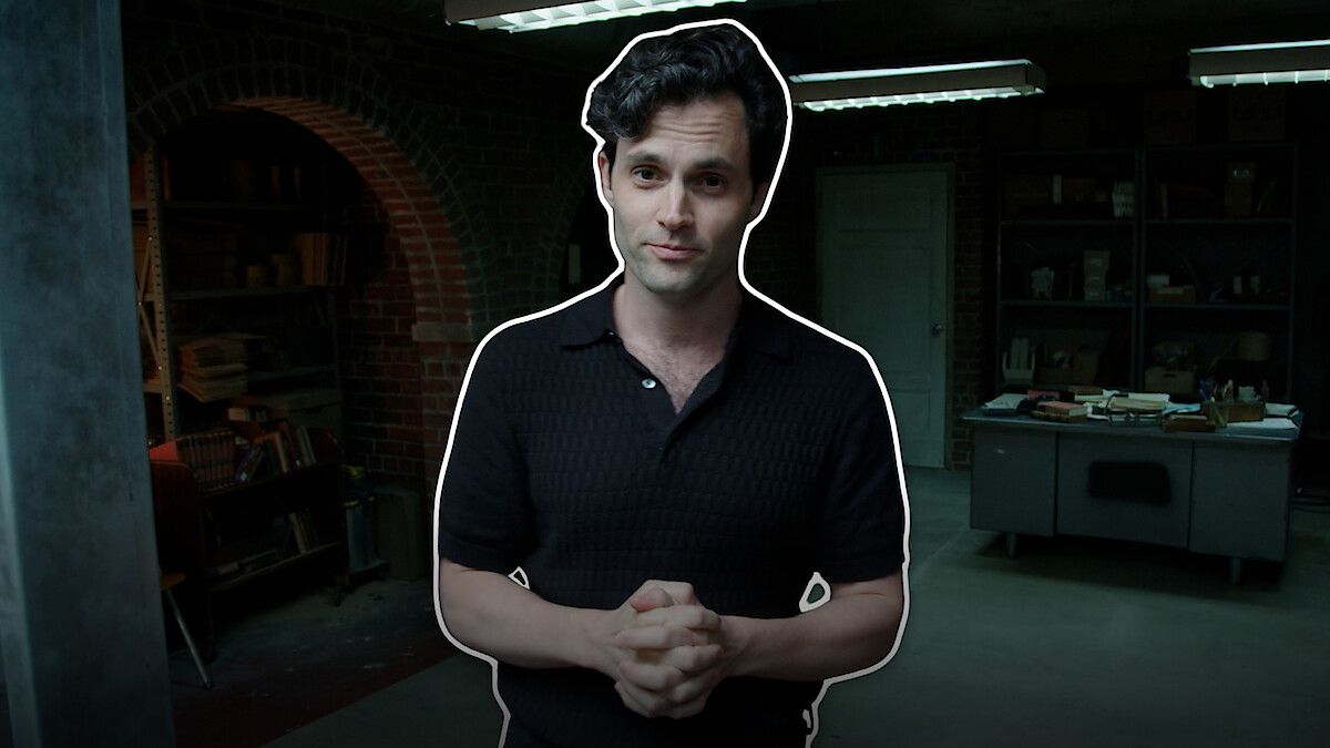 Penn Badgley, wearing a dark shirt, stands in a dimly lit room with brick walls and shelves. He has his hands clasped and is looking at the camera.