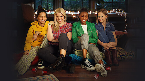 Four young women sit on the floor together in a cozy room with string lights and large windows, smiling and relaxed, surrounded by pillows and cups, suggesting a casual indoor gathering or hangout.