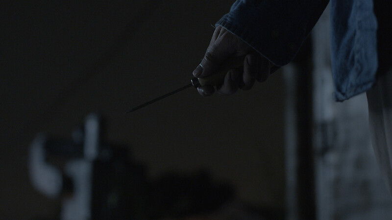 A person holding a knife in their right hand at night, wearing a dark blue jacket. The background is dimly lit, creating a suspenseful or tense atmosphere.