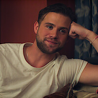 Leo Woodall wearing a white t-shirt, relaxes on a couch with his arm resting on a cushion in a cozy indoor setting with patterned curtains in the background.