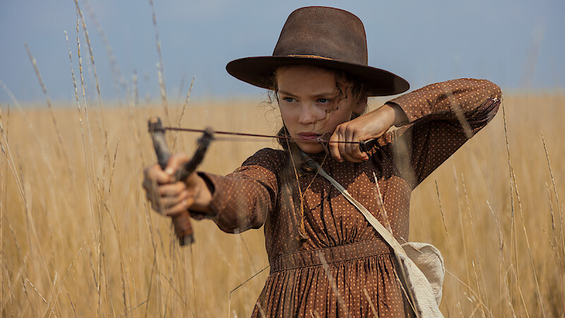 Girl in a brown dress and wide-brim hat aims a slingshot in a field of tall, dry grass under a clear sky, looking focused and determined.