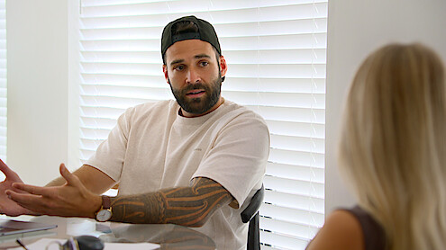 Alex wearing a cap sits at a table talking to a blonde woman in a bright room with white blinds, appearing engaged in conversation.