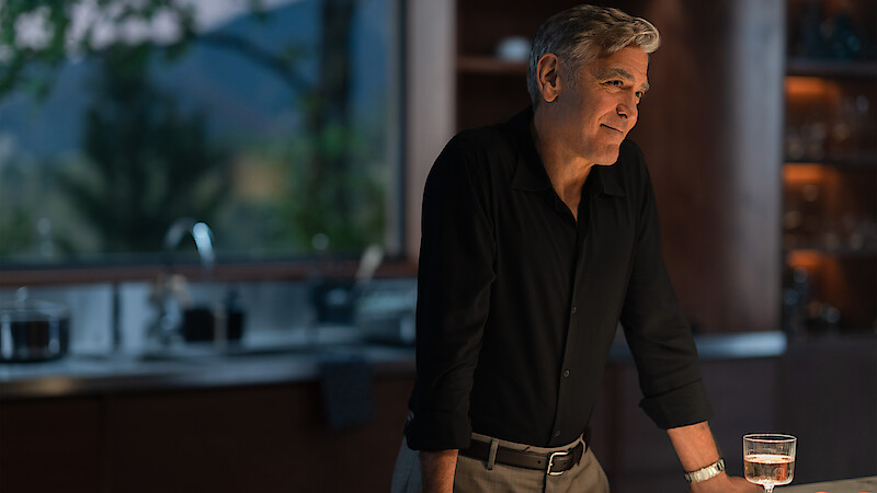 Jay Kelly (George Clooney) stands smiling at the countertop in his vast kitchen while Peter Schneider (Jim Broadbent) makes him a sandwich off camera.