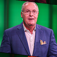 An older man with glasses in a blue blazer and white shirt stands in front of a bright green background, possibly on a studio set.