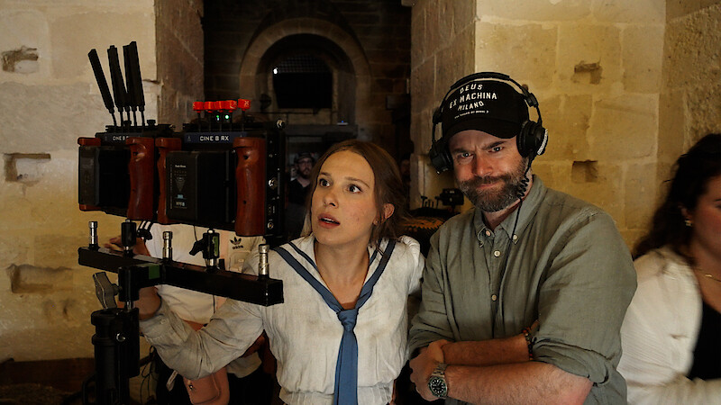 A woman in costume and a man with headphones stand by a camera rig in a stone hallway on a film set, reviewing footage together.