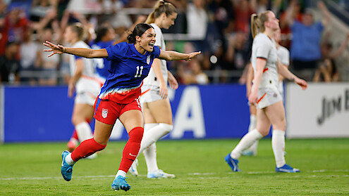 Soccer player in blue jersey celebrates with arms outstretched on a stadium field, surrounded by other players in white uniforms and a cheering crowd in the background during a match.