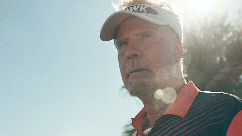 A man wearing a sports visor and striped shirt stands outdoors in the sunlight, with palm trees and blue sky in the background, giving a serious expression.