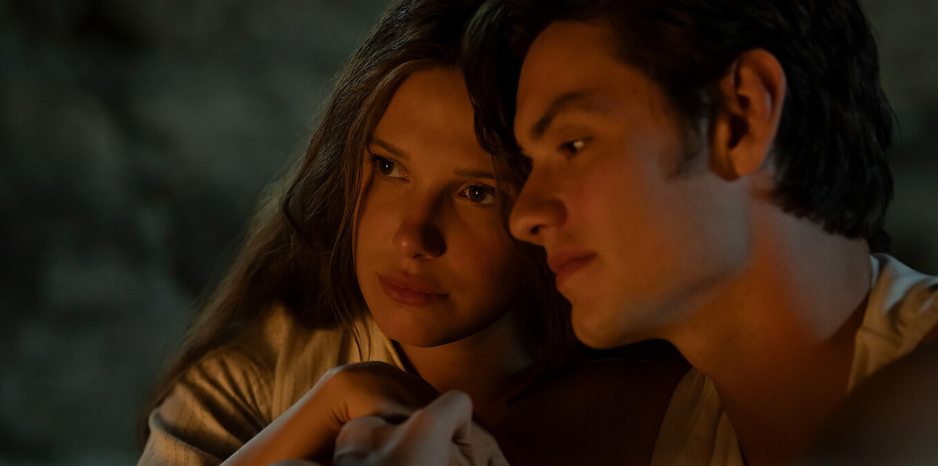 Two young people sit closely together in a dimly lit, cozy setting, with warm light illuminating their faces, suggesting an intimate or reflective moment.