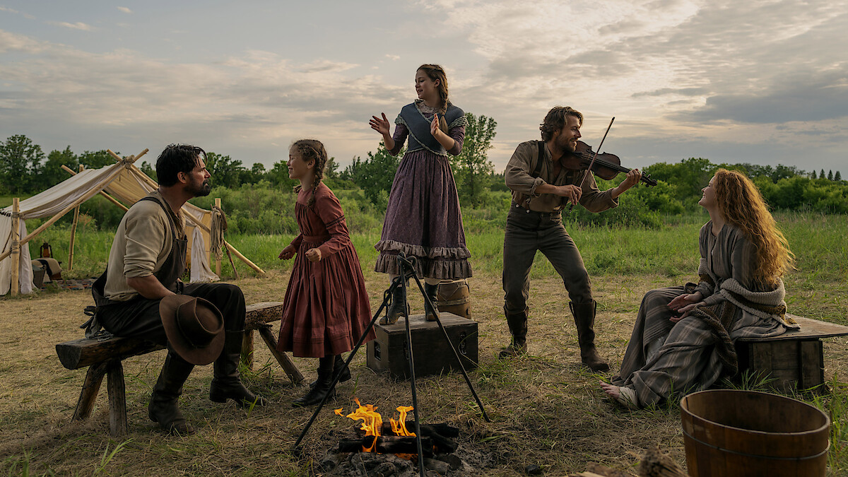 Five people in pioneer-era clothing gather around a campfire in an open field with tents. One plays a violin, others sing or listen, creating a lively, outdoor, historical family campsite scene at sunset.