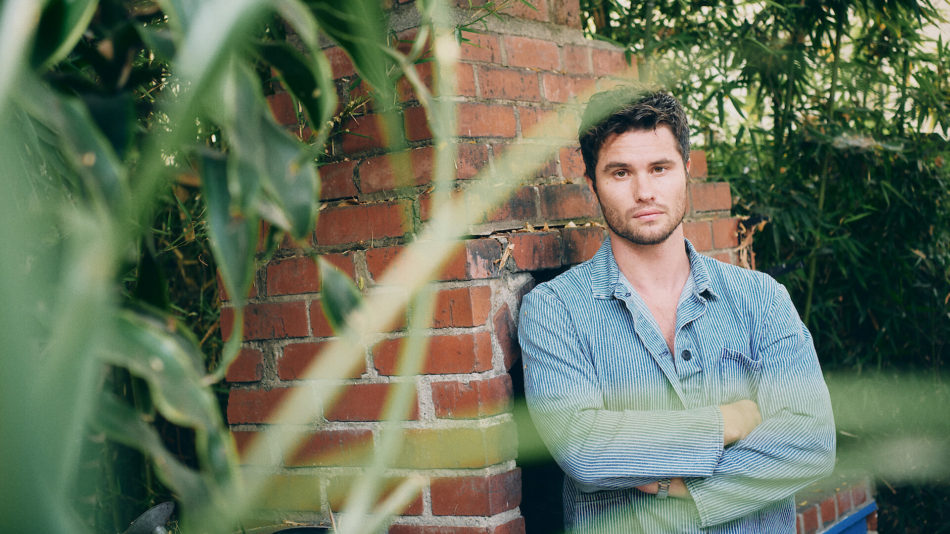 A man in a light denim shirt stands with arms crossed in front of a brick wall outdoors, surrounded by green plants and bamboo. The setting is natural and relaxed, with leaves partially obscuring the foreground.