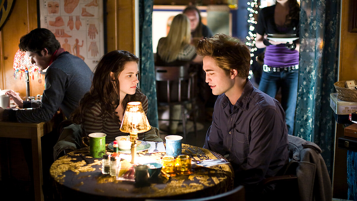 A young woman and man sit at a small round table with drinks and a lamp between them in a cozy, warmly-lit café, talking closely while other people sit and walk in the background.