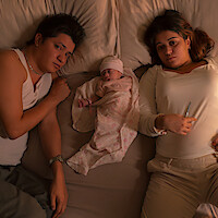 A man and woman lie on a bed with a newborn baby swaddled between them. The setting is a bedroom with warm lighting, suggesting a family moment. The woman holds a thermometer, possibly indicating concern for the baby's health.
