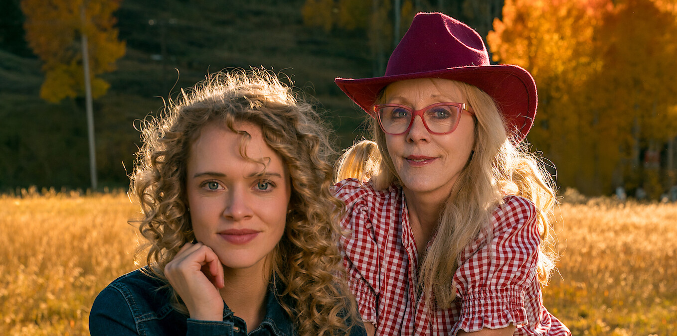 Two women in a field with autumn trees; one with curly hair wearing denim, the other in glasses, a red hat, and checkered shirt. Warm sunlight, outdoors, fall setting.