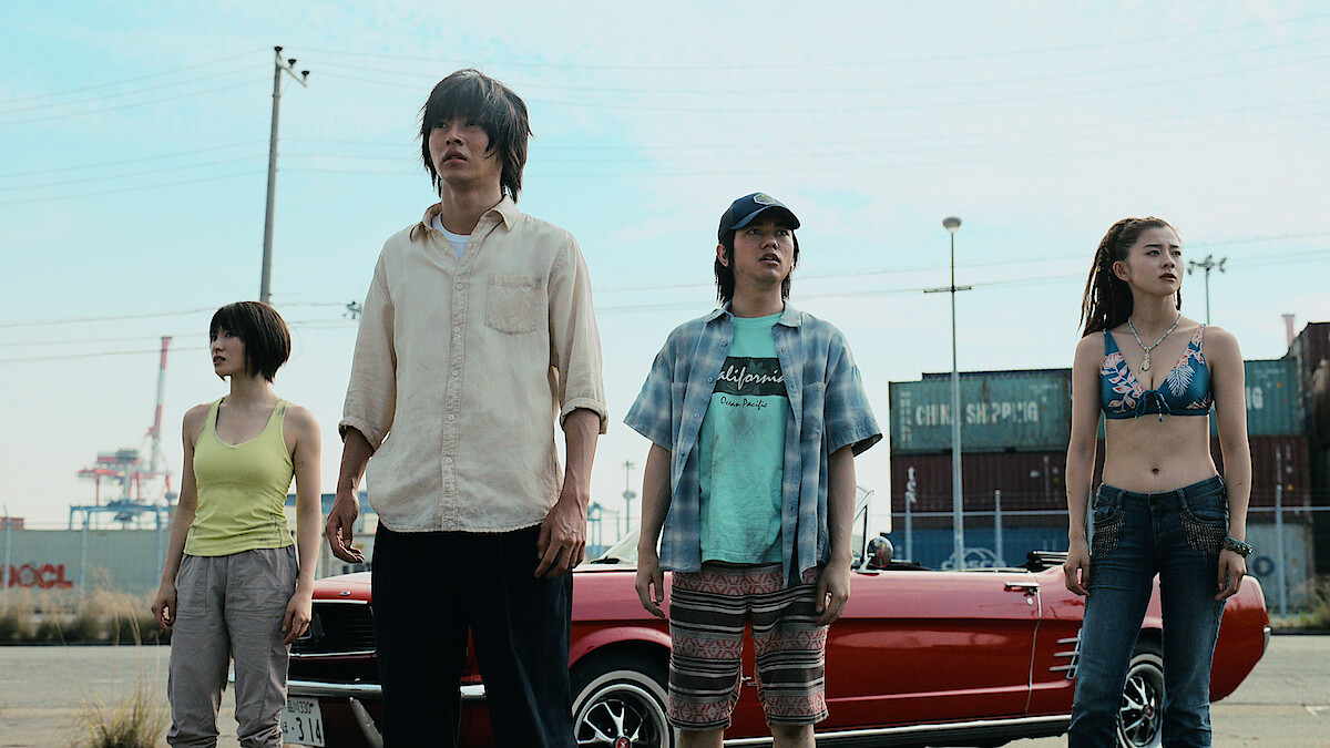Four people stand in front of a red convertible car in an industrial area with shipping containers and power lines visible in the background, under a clear sky.