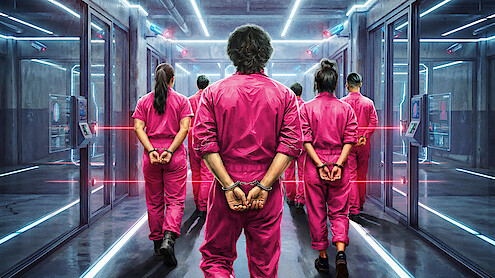 Several people in pink jumpsuits, hands cuffed behind their backs, are being escorted down a futuristic, neon-lit corridor lined with glass walls and security panels.